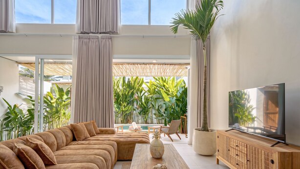 A modern living room with a large sectional sofa, a wooden coffee table, a TV on a wooden cabinet, and a view of a lush garden through large windows. - Villa Pia Pererenan (Munggu)