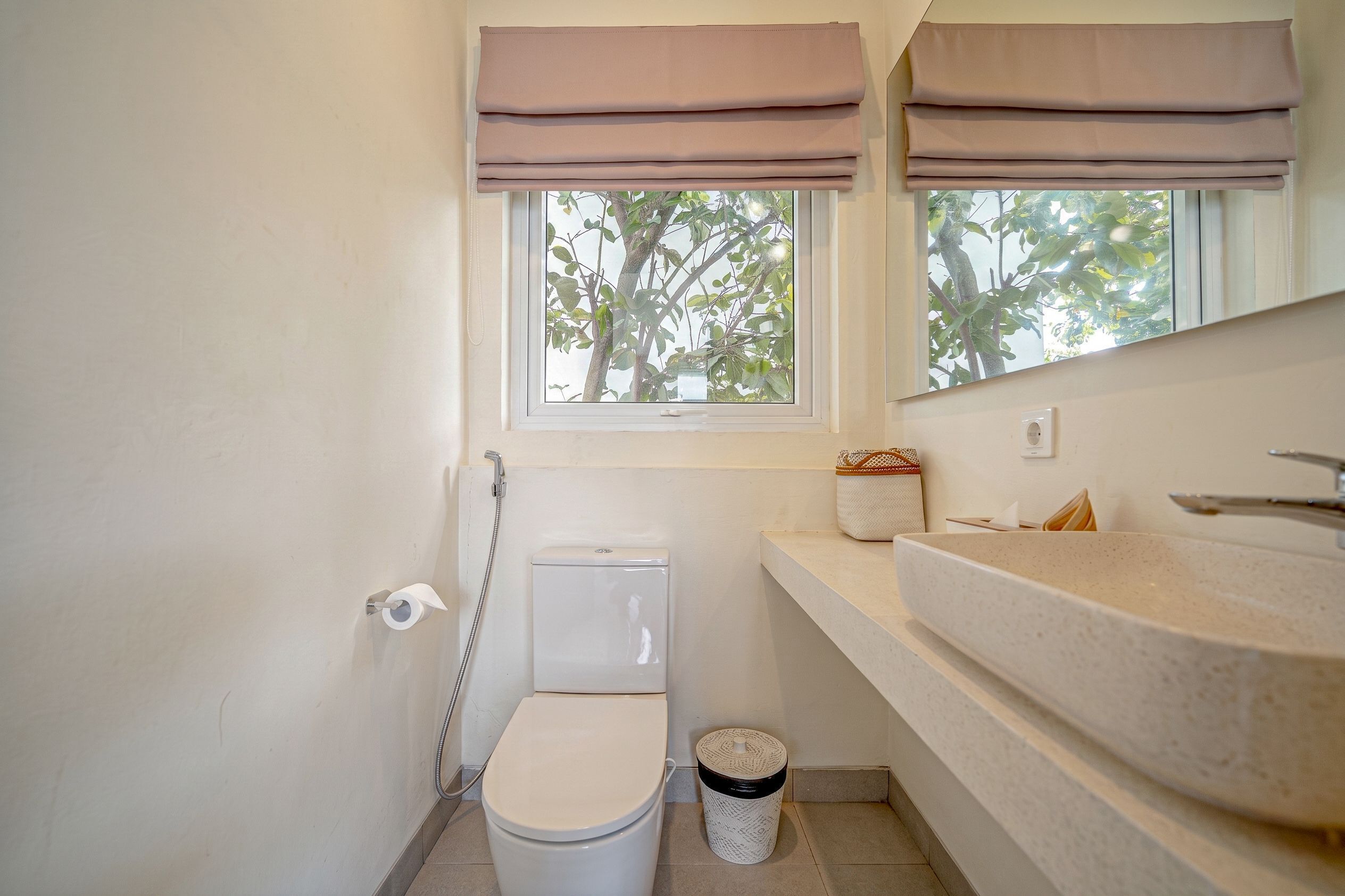 A bathroom with a toilet, bidet, sink, and window with a view of trees.