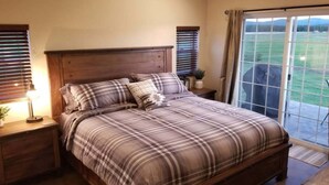 Un dormitorio con una cama grande, cabecera de madera, dos mesitas de noche con lámparas y vistas a un paisaje verde a través de una puerta corredera de vidrio.