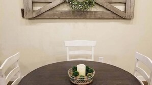Una mesa redonda con mantel marrón oscuro, rodeada por cuatro sillas blancas. Sobre la mesa, una decoración de pared de madera con un diseño de cruz y un pequeño arreglo floral.