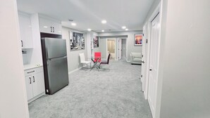 Een moderne keuken met witte kastjes en een roestvrijstalen koelkast. Er is een eethoek met een glazen tafel en stoelen. De vloer is bekleed met een grijs vloerkleed.
