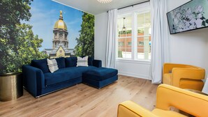Un salon avec un canapé d’angle bleu, un fauteuil jaune, une grande fenêtre avec des rideaux blancs et une fresque représentant un bâtiment surmonté d’un dôme.