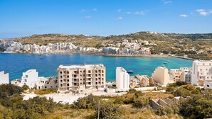 A coastal town with buildings near the water, a clear sky, and a hilly landscape in the background.