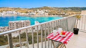 A balcony with a table set for two, overlooking a coastal town with buildings and a clear blue sea.