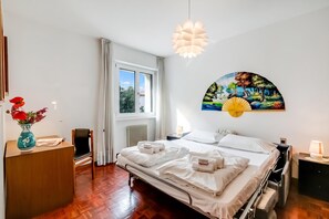 A hotel room with a bed, bedside tables, a desk, a chair, a radiator, a window with curtains, and a wall-mounted artwork. - Dora - Riva del Garda Apartment with Terrace, Riva del Garda, Italy (Riva del Garda)