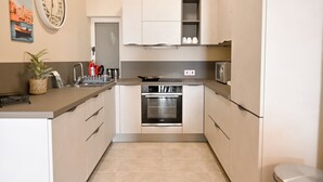 A compact kitchen with a built-in oven, microwave, and a small sink. There is a potted plant on the counter and a decorative wall clock. - Sea & Valley Views Żebbuġ Gozo, Żebbuġ Gozo, Malta (Żebbuġ Gozo)