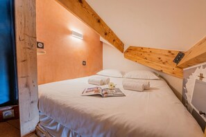 Camera da letto con soffitto in legno, un letto con lenzuola bianche, una rivista sul letto e una lampada fissata al muro.