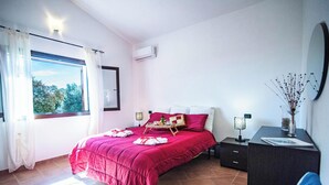 A bedroom with a large window, a bed with a red bedspread, a bedside table with a lamp, and a dresser.