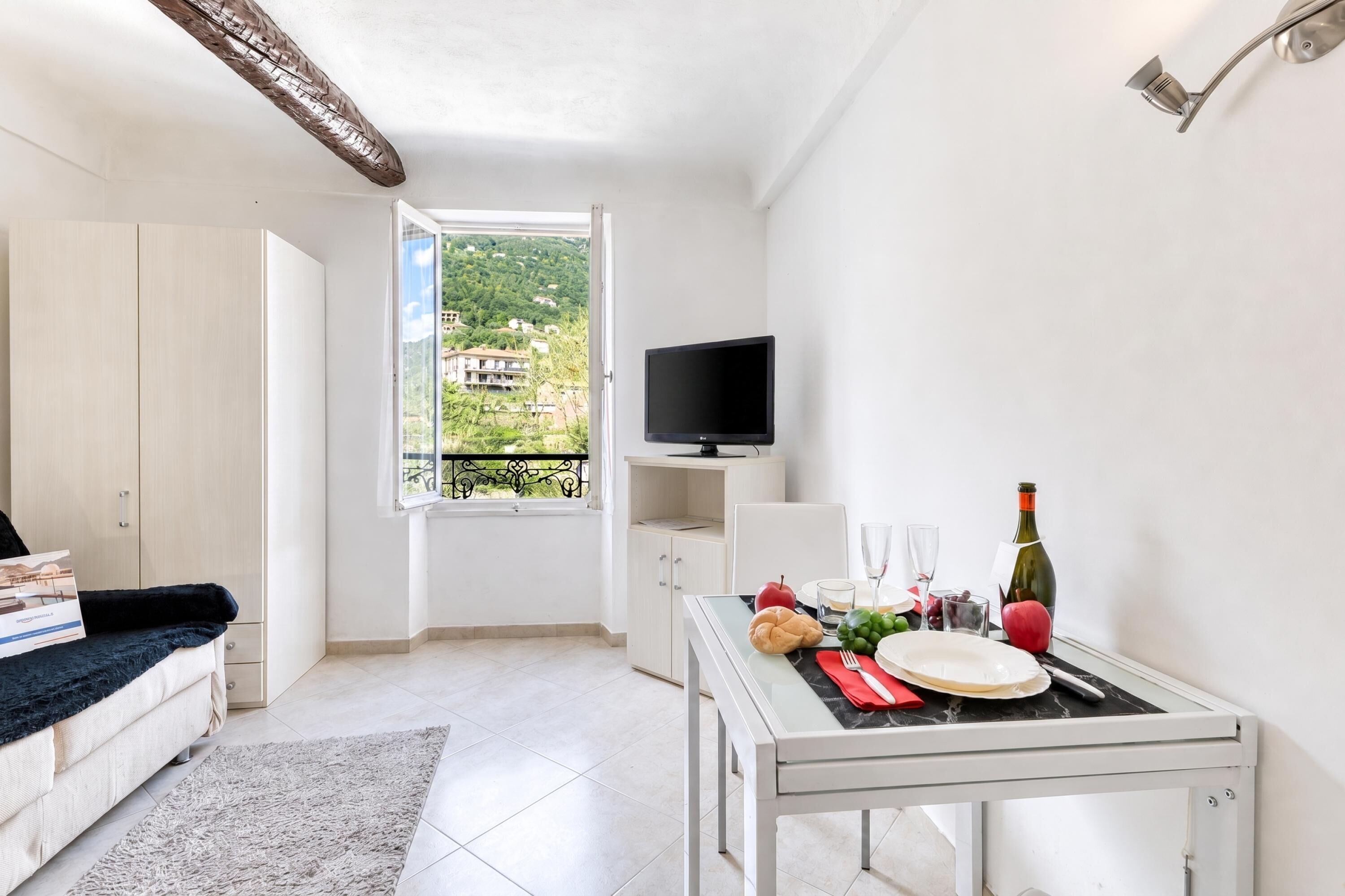 Una sala de estar moderna con un sofá, una mesa de comedor para dos personas, un televisor y un balcón con vistas a una zona residencial.