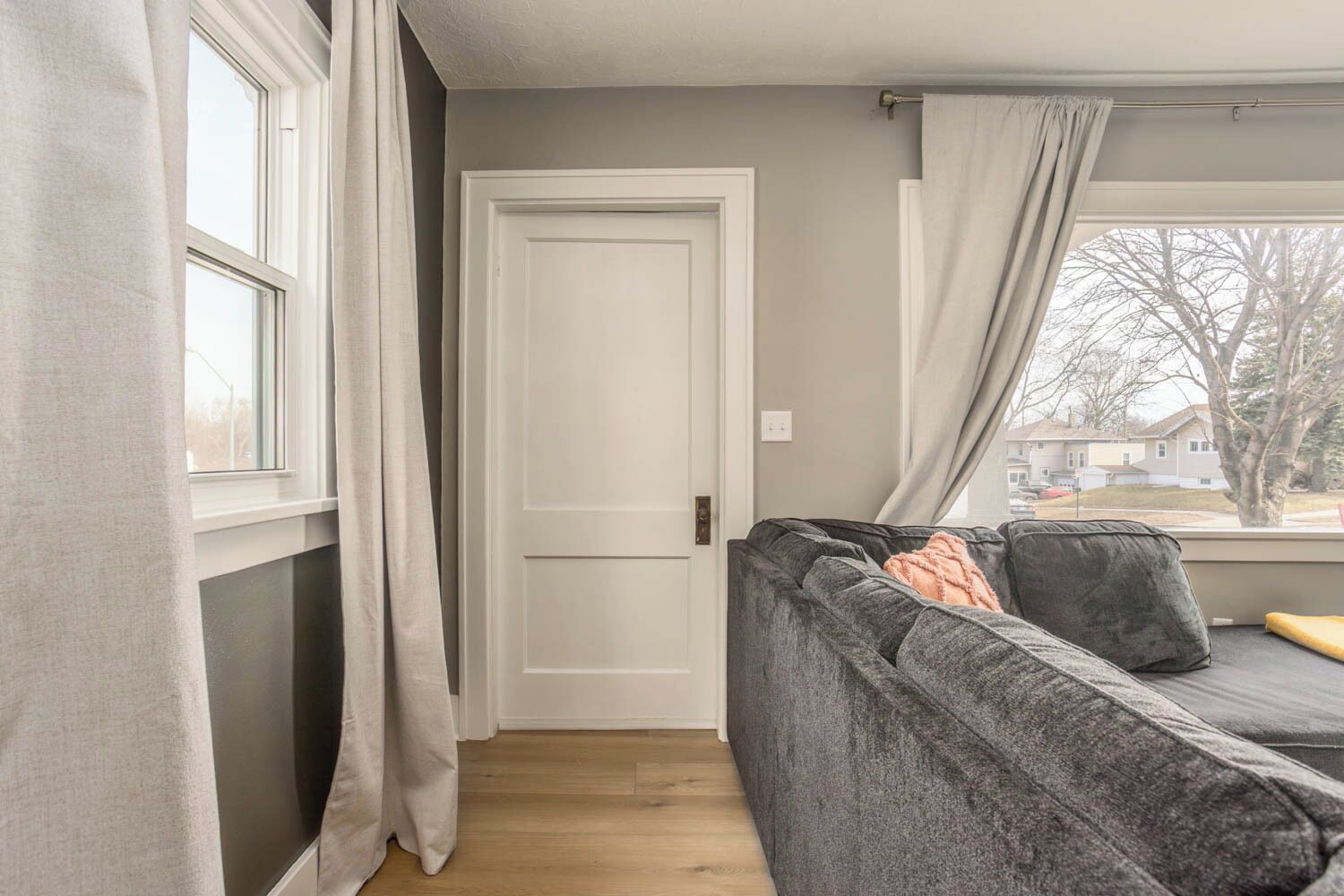 A room with a grey sofa, a closed white door, and a window with curtains.