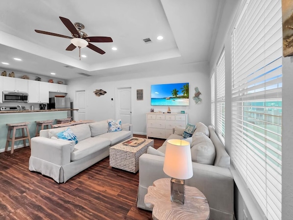 A modern living room with a sofa, coffee table, and a lamp. There is a kitchen area with cabinets and a ceiling fan.