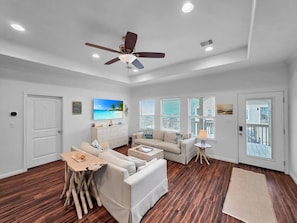 A spacious living room with a ceiling fan, a TV, and a view of the ocean through large windows. - Steps to the Beach • Fenced Yard • Sleeps 8 with Fully Stocked Kitchen (Bolivar Peninsula)