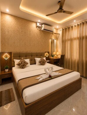 A hotel room with a large bed, a ceiling fan, an air conditioning unit, a desk with a chair, and a large mirror. - Stay Purvi Ganga (VARANASI)