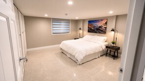A bedroom with a bed, bedside tables, a window with blinds, a painting on the wall, and a door.