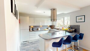 Uma cozinha moderna com armários brancos, uma ilha central com banquetas, um exaustor de aço inoxidável e uma janela que oferece luz natural.