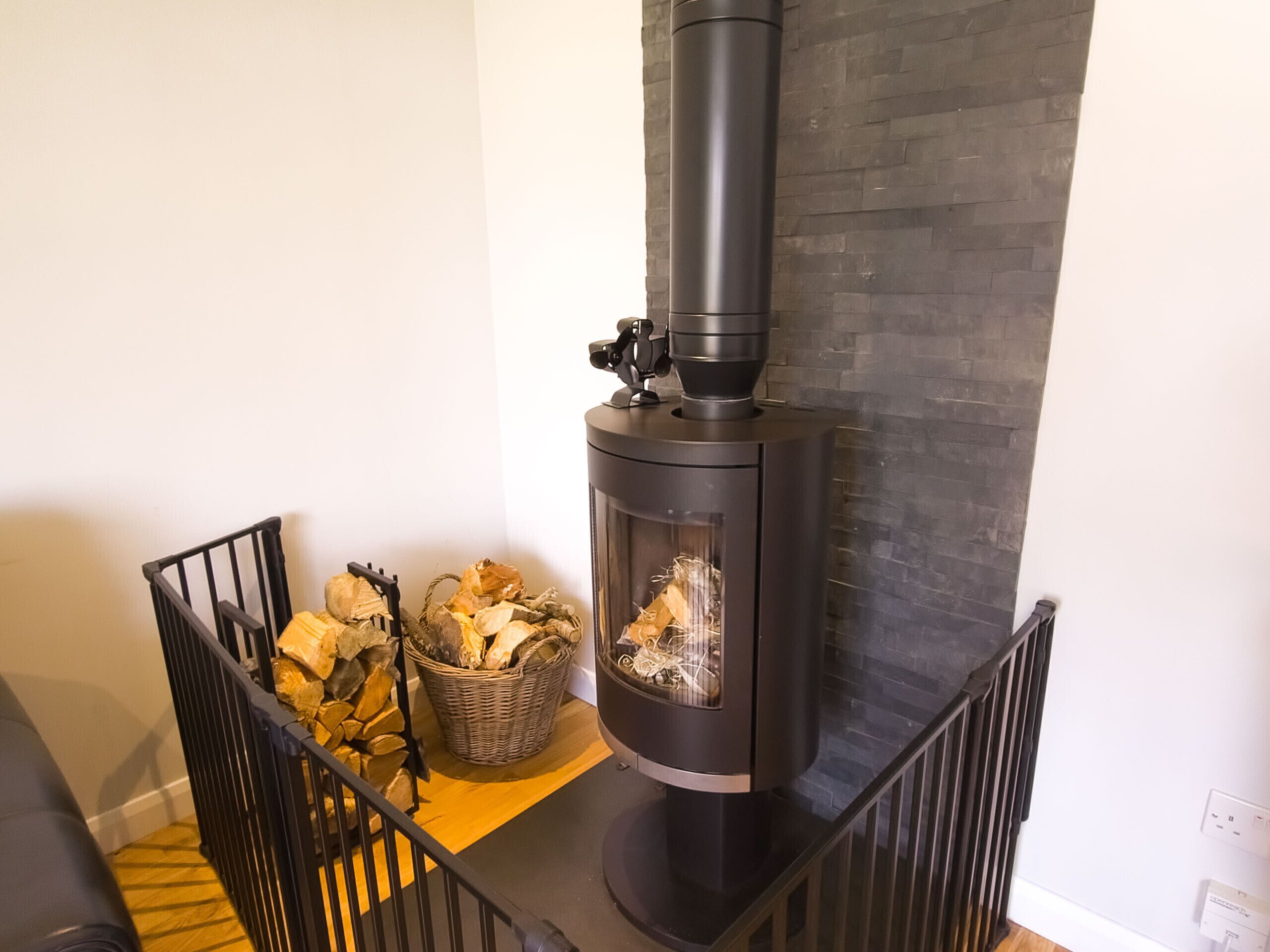 A modern living room with a black wood-burning stove, a wicker basket filled with firewood, and a black metal railing.