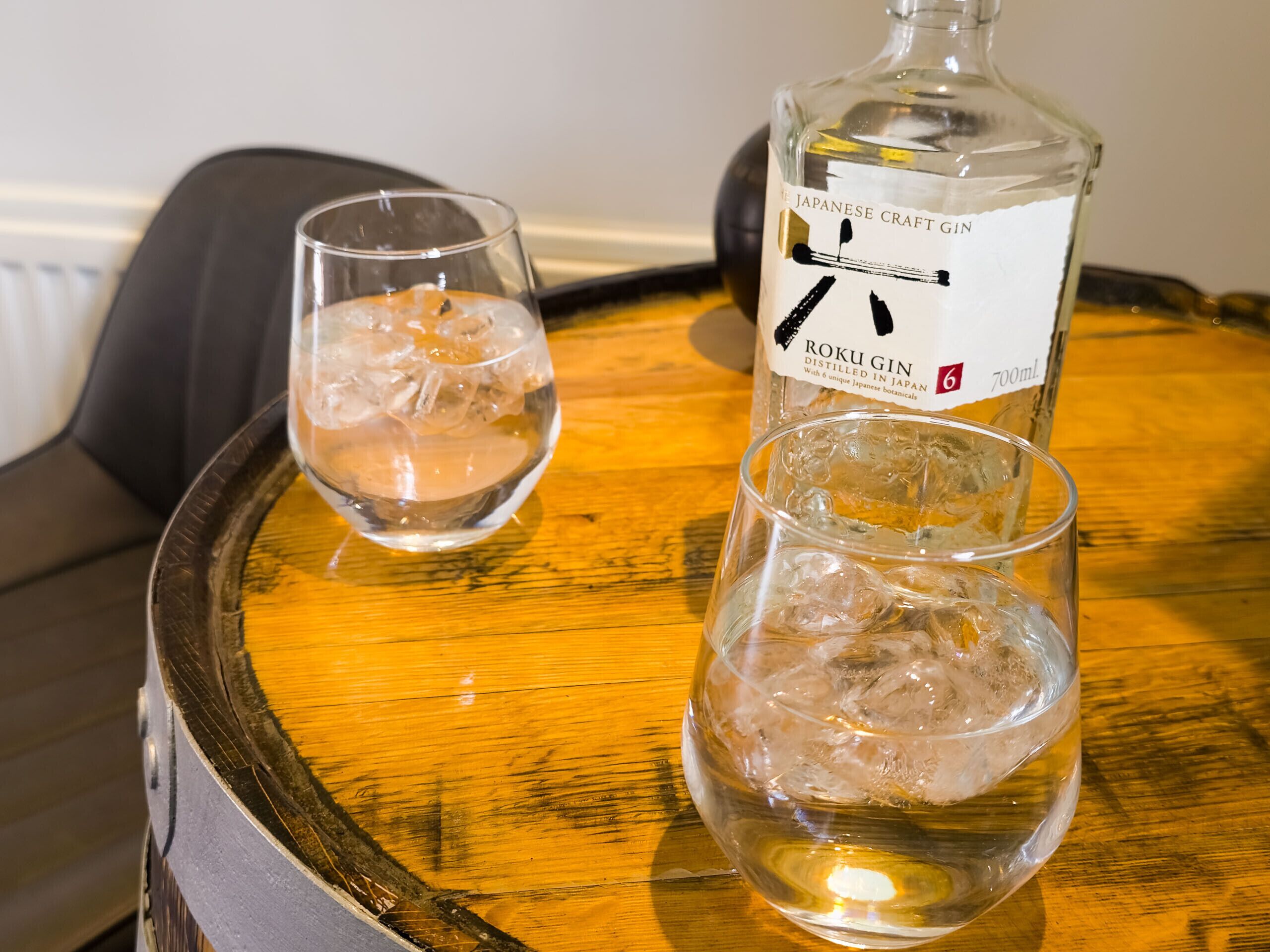 Two glasses of a clear liquid with ice cubes on a wooden tray, and a bottle labeled "Roku Gin" on a table.
