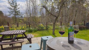Um pátio externo com mesas e cadeiras de madeira, um barril e uma garrafa de vinho com um copo sobre uma mesa.