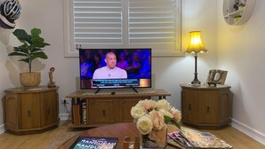 A living room with a television, wooden cabinets, a potted plant, a vase with flowers, and a magazine on the coffee table. -  Private Central Guesthouse (Dudley Park)