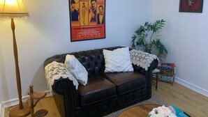 A living room with a dark leather sofa, a lace throw, a poster on the wall, a lamp, and a potted plant. -  Private Central Guesthouse (Dudley Park)
