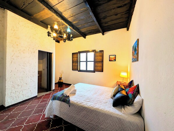 Un dormitorio con cama, una araña, una ventana con contraventanas y un cuadro en la pared.