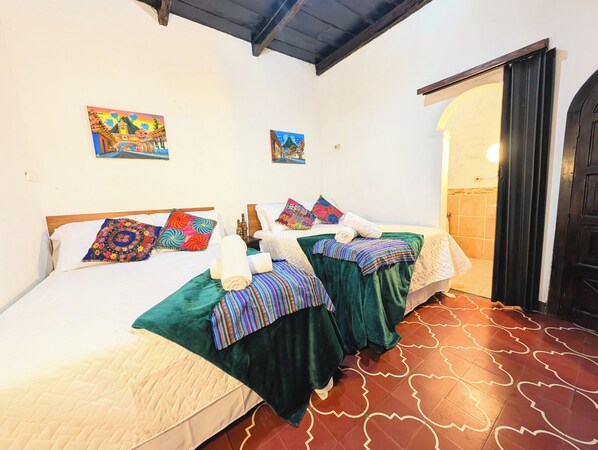 A room with two beds, each with colorful bedding and pillows, a tiled floor, and two paintings on the walls. - Casa Quetzalli Los Nazarenos (Antigua Guatemala)