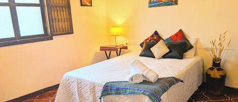 A neatly made bed with a quilted bedspread, pillows, and a rolled towel. There is a small table with a lamp beside the bed, and a vase with dried plants on the floor.