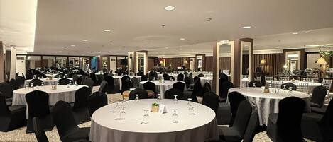 A large banquet hall with round tables, black chairs, and a carpeted floor.