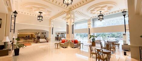 Un amplio vestíbulo con techos altos, columnas ornamentadas y candelabros. Hay zonas de estar con sofás y mesas, y ventanas grandes que ofrecen luz natural.