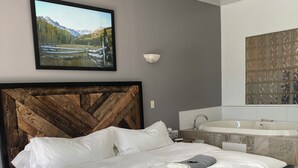 A modern bedroom with a wooden headboard, a large bed with white linens, a mounted TV, a bathroom with a bathtub, and a decorative wall panel. - Pikes Peak Paradise Bed and Breakfast (Woodland Park)