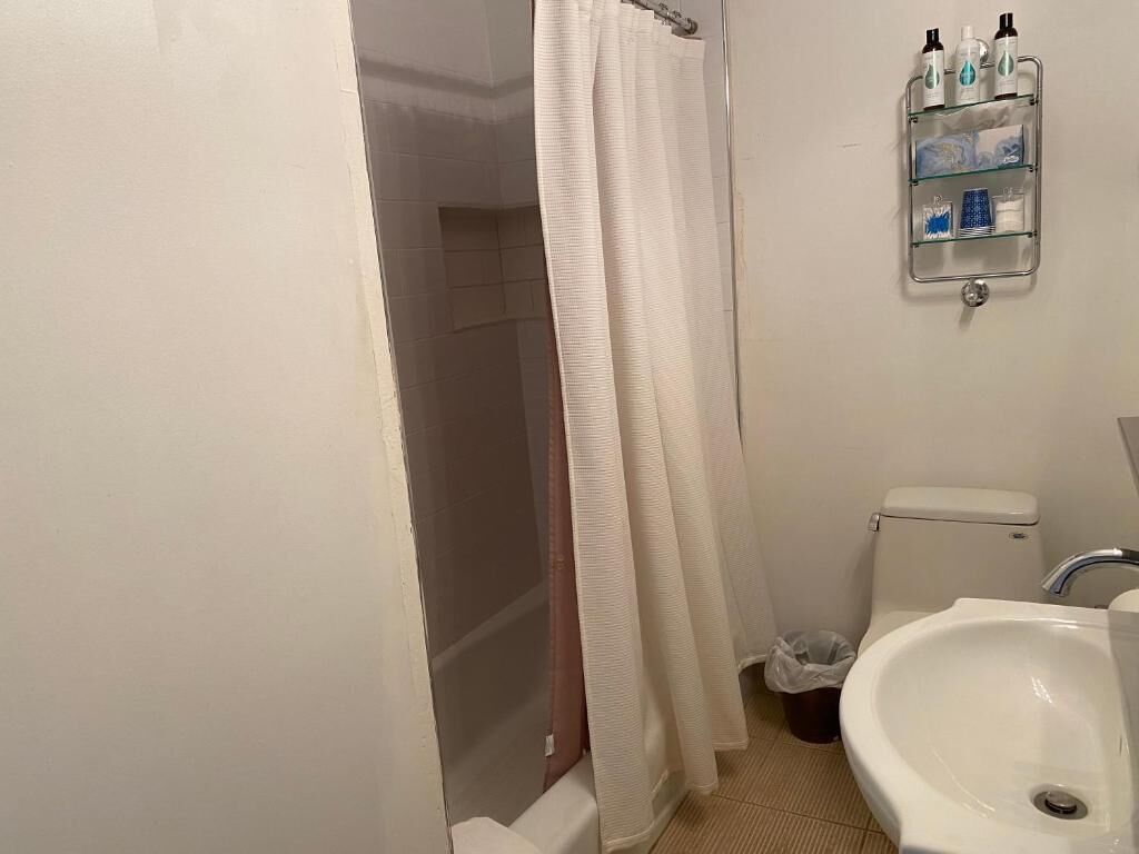 A bathroom with a shower, toilet, and sink. There is a shower curtain, a glass shelf with toiletries, and a trash bin.