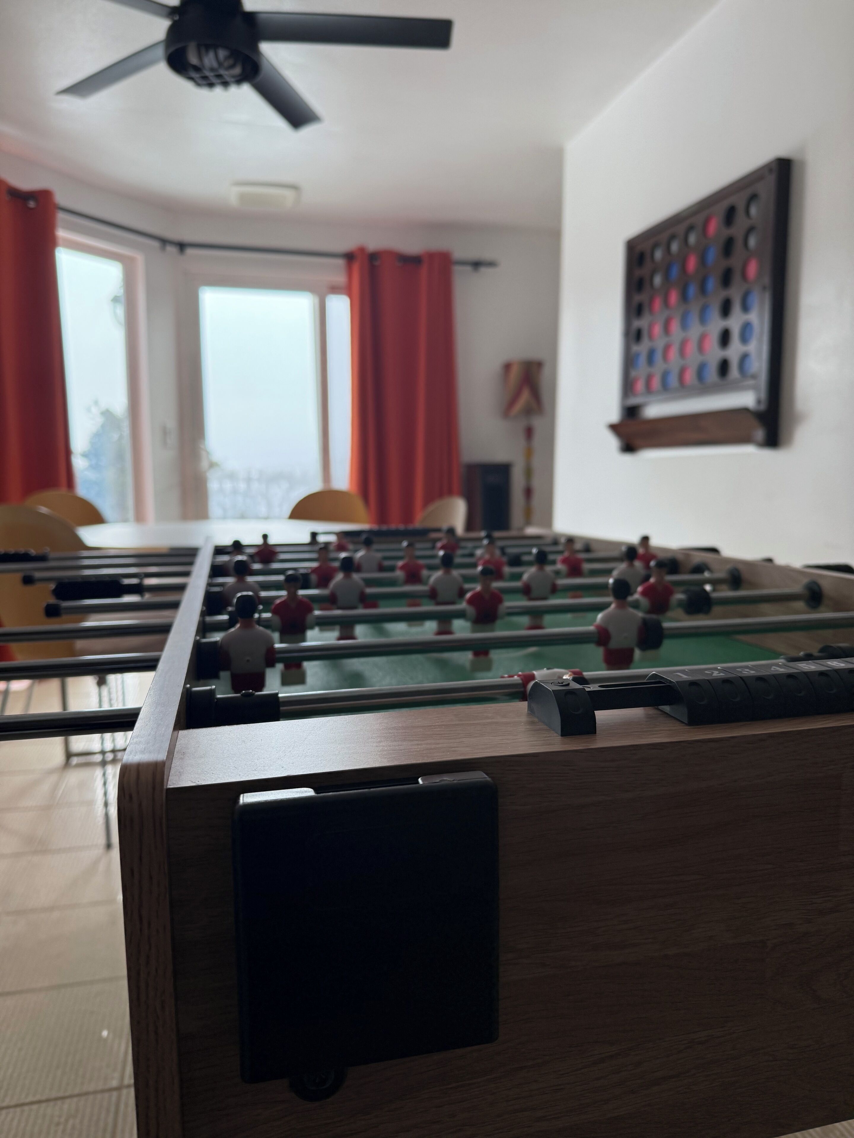Una mesa de fútbol de mesa en una habitación con ventanas, cortinas rojas y un ventilador de techo.