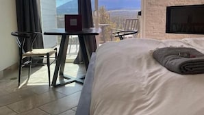 A hotel room with a large bed, a desk, a chair, and a TV. There is a view of mountains through the window. - Pikes Peak Paradise Bed and Breakfast (Woodland Park)