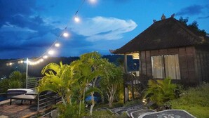 Un edificio con techo de paja y balcón, rodeado de plantas tropicales y una zona de piscina con sillas de descanso.