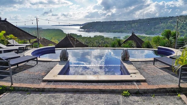 Una piscina con dos estatuas de piedra, rodeada de sillones de descanso y una vista panorámica de un paisaje costero.