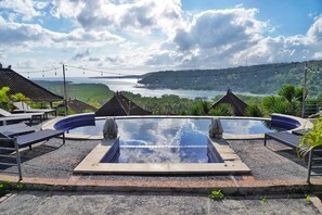 A swimming pool with two stone statues, surrounded by lounge chairs and a scenic view of a coastal landscape.
