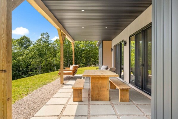 A covered outdoor patio with a wooden table and benches, surrounded by a gravel path and greenery.