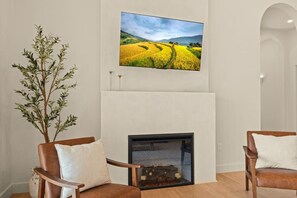 A living room with a fireplace, two chairs, a large flat-screen TV mounted above the fireplace, and a tall plant.
