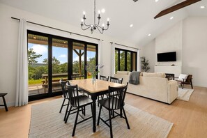 A modern living room with a dining area, large windows, a flat-screen TV, and a fireplace.