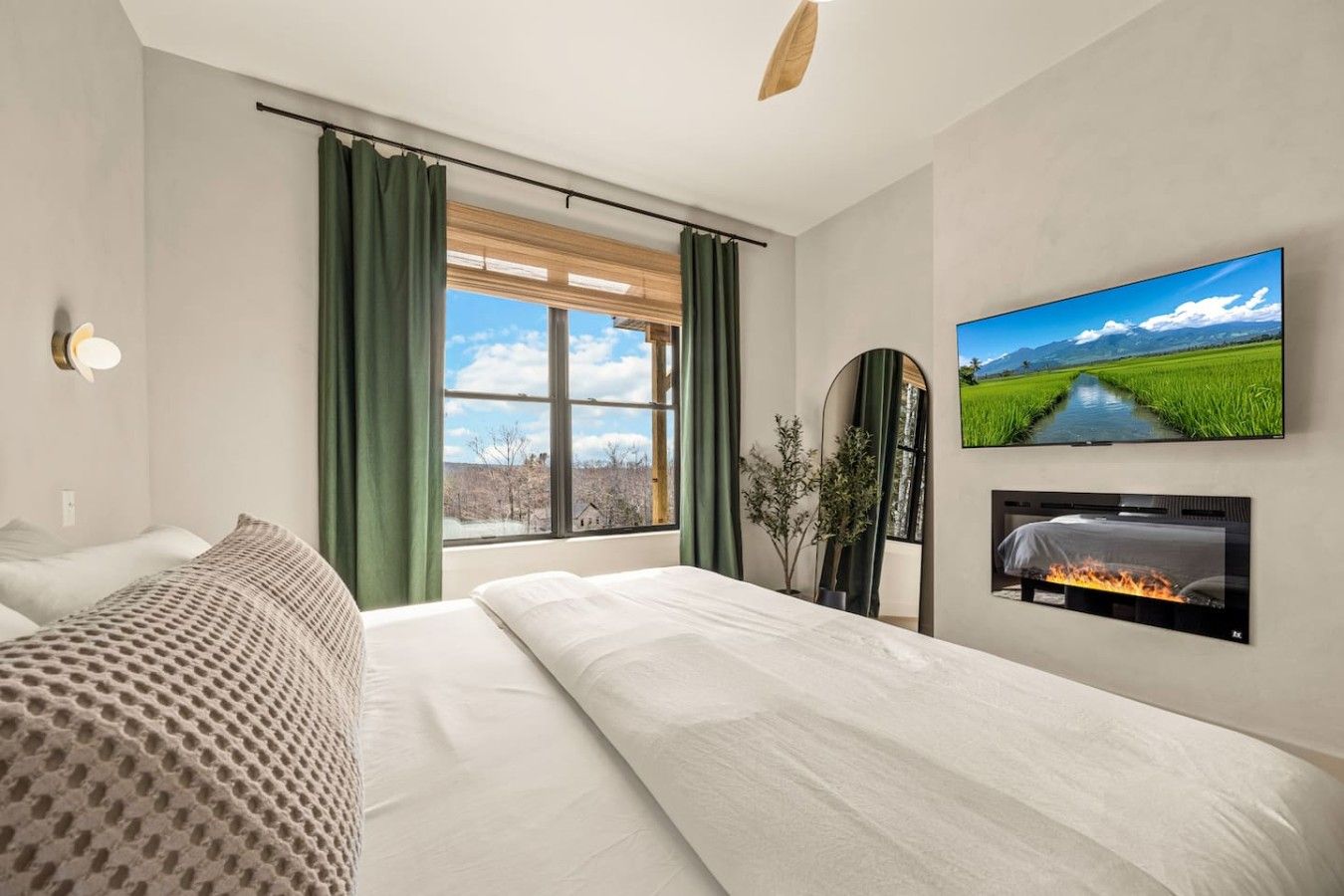 A hotel room with a large bed, a flat-screen TV mounted on the wall, a fireplace, and a window with curtains.