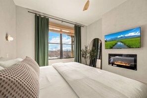 A hotel room with a large bed, a flat-screen TV mounted on the wall, a fireplace, and a window with curtains.