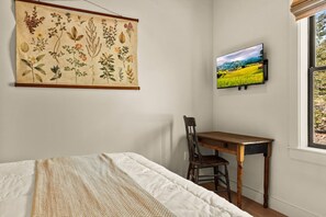 Quarto com cama, escrivaninha e cadeira, uma grande ilustração botânica na parede e uma TV exibindo uma paisagem.