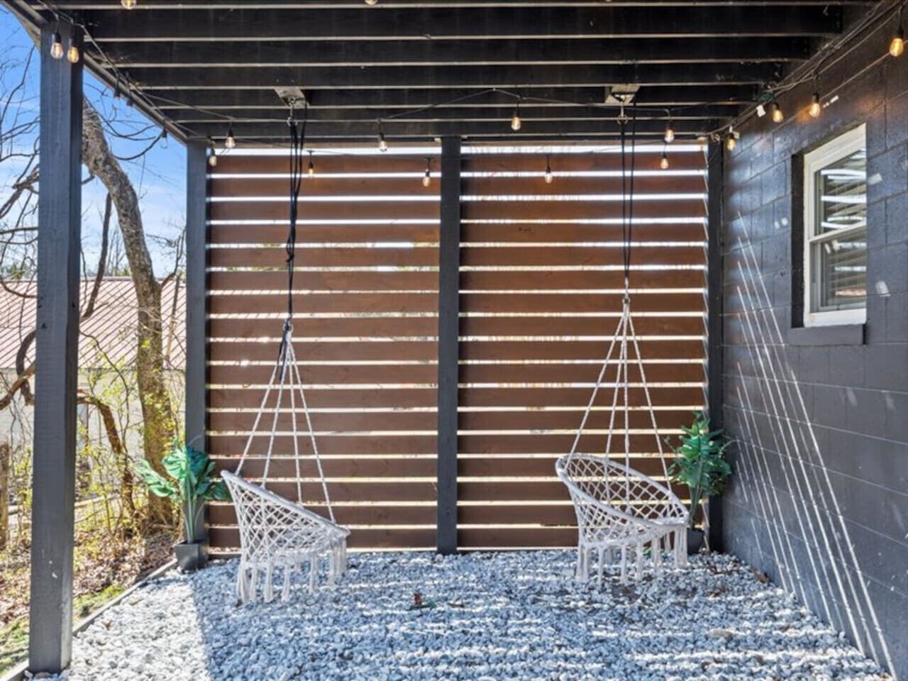 Un espace extérieur abrité avec deux chaises blanches et un hamac, un mur en bois à lattes et un sol recouvert de gravier.
