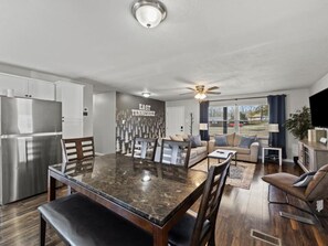 A modern kitchen with a large island, a dining area with a glass-top table, and a living room with a sofa and TV.