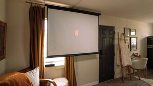 Ett mysigt vardagsrum med en stor tv-skärm, en soffa, en fåtölj, ett litet bord och en dörr.
