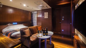 A modern hotel room with a wooden floor, a leather sofa, a small round table with wine glasses and bottles, a bed with a large pillow, and a TV on the right wall.