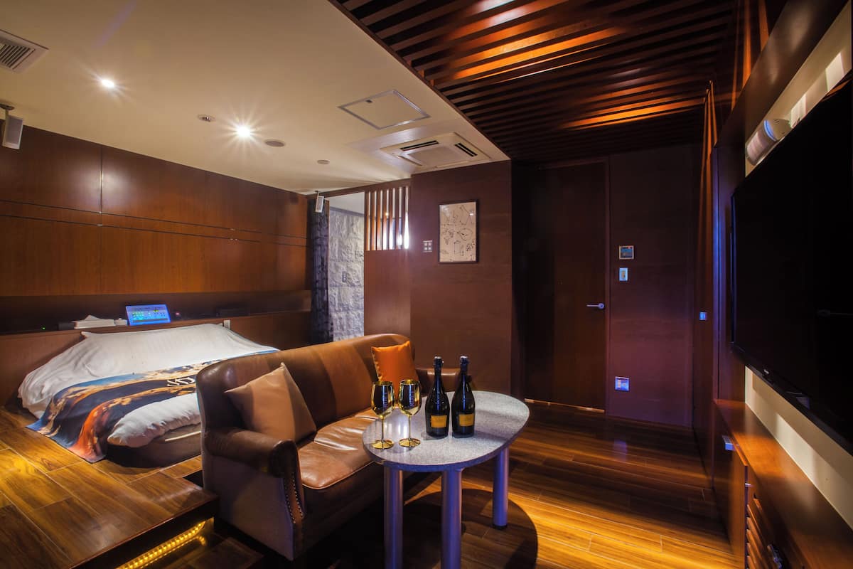 A modern hotel room with a wooden floor, a leather sofa, a small round table with wine glasses and bottles, a bed with a large pillow, and a TV on the right wall.