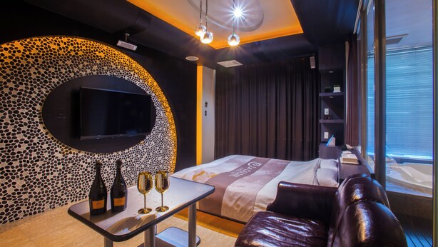 A modern hotel room with a curved wall mosaic, a flat-screen TV, a bed with a decorative headboard, a leather sofa, and a small table with wine bottles and glasses.