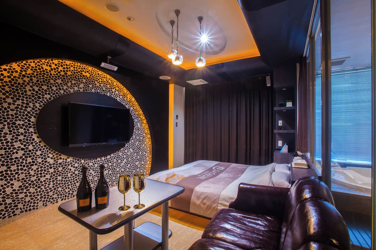 A modern hotel room with a curved wall mosaic, a flat-screen TV, a bed with a decorative headboard, a leather sofa, and a small table with wine bottles and glasses.
