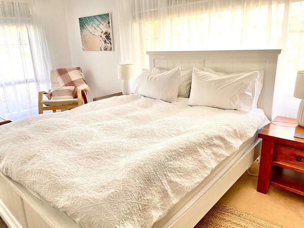 Una cama bien hecha con una colcha blanca, una mesita de noche de madera con una lámpara, y una silla con una manta de cuadros.